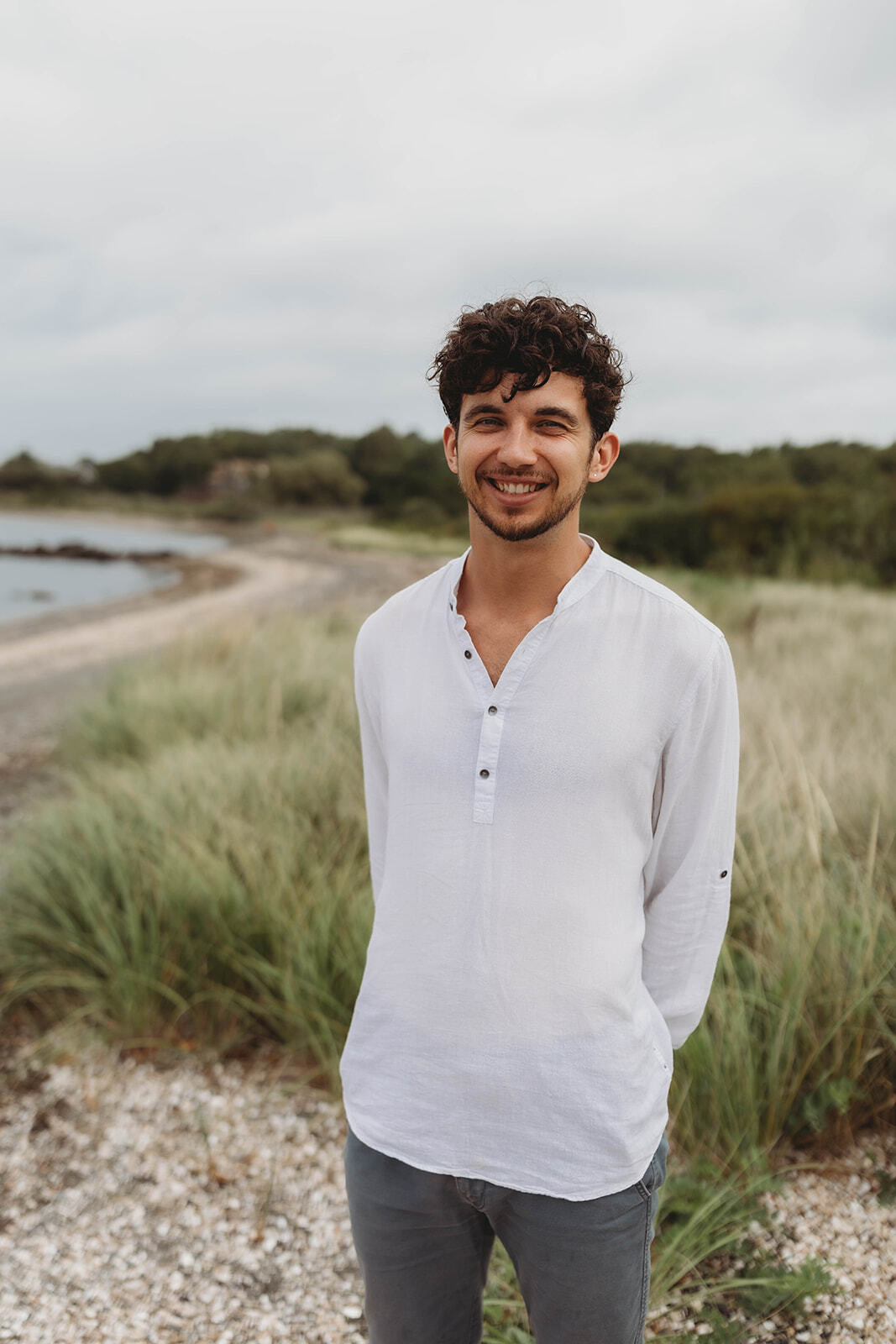 Dr. Ben Thompson, AuD standing outdoors in a white linen shirt with a blurred dune and treeline behind him
